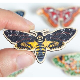 wooden pin with a print of a deaths head hawkmoth. Held on the left between a finger and thumb. Out of focus moth pins in the background.