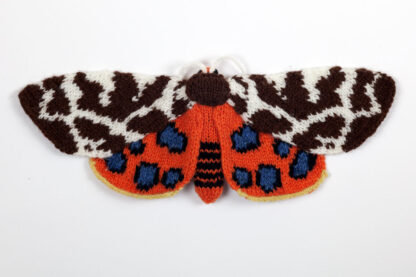 A knitted moth on white background. The Upper wings are a patchwork of brown and white. The under wings are bright orange with 5 blue and black spots